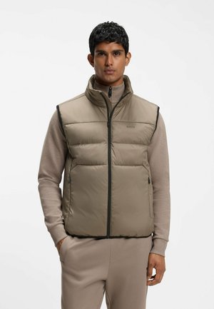 Mann trägt beige gesteppte Weste über passendem Reißverschluss-Pullover und Hose, steht mit einer Hand in der Tasche, neutraler Hintergrund.