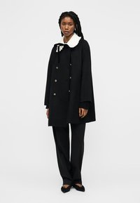 Γυναίκα με πλεγμένα μαλλιά που φοράει μαύρο διπλό παλτό με λευκό γιακά, μαύρο παντελόνι και μαύρες μπαλαρίνες, στέκεται μπροστά σε λευκό φόντο.