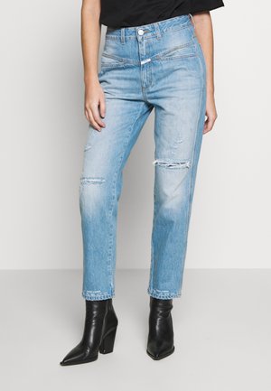 Osoba w jasnoniebieskich podartych dżinsach i czarnych szpiczastych botkach na obcasie, stojąca na tle jednolitego jasnego tła.