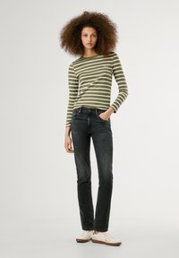 Olīvzaļganā svītrainā garais piedurkņu tops ar apaļu kakla izgriezumu, apvienots ar tumšām džinsām. Modelis nēsā gaišus apavus, rokas kabatās, ar lokainiem matiem.