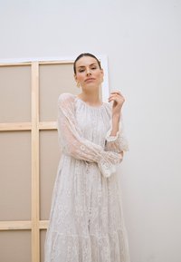 Femme aux cheveux tirés en arrière, portant une longue robe blanche en dentelle et des boucles d'oreilles dorées, debout à l'intérieur devant un mur uni et un décor en cadre en bois.