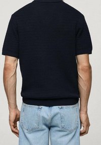 Homme portant un t-shirt à manches courtes texturé noir et un jean en denim bleu clair, vu de dos sur un fond uni.