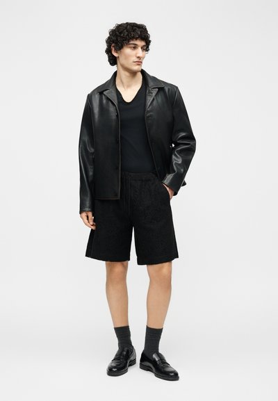 Νεαρός με σγουρά μαλλιά που φοράει μαύρο δερμάτινο μπουφάν, μαύρο μπλουζάκι με V λαιμόκοψη, μαύρο σορτς με υφή, γκρι κάλτσες και μαύρα δερμάτινα παπούτσια.