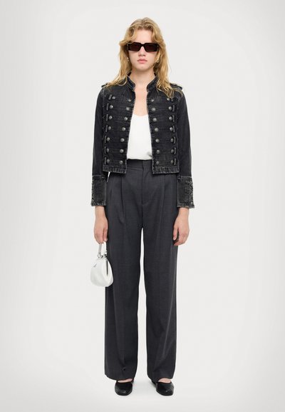 Šviesiaplaukis asmuo dėvi juodą užsegamą švarką, baltą palaidinę, tamsias plačias kelnes, juodus batelius, akinius nuo saulės ir laiko mažą baltą rankinę.