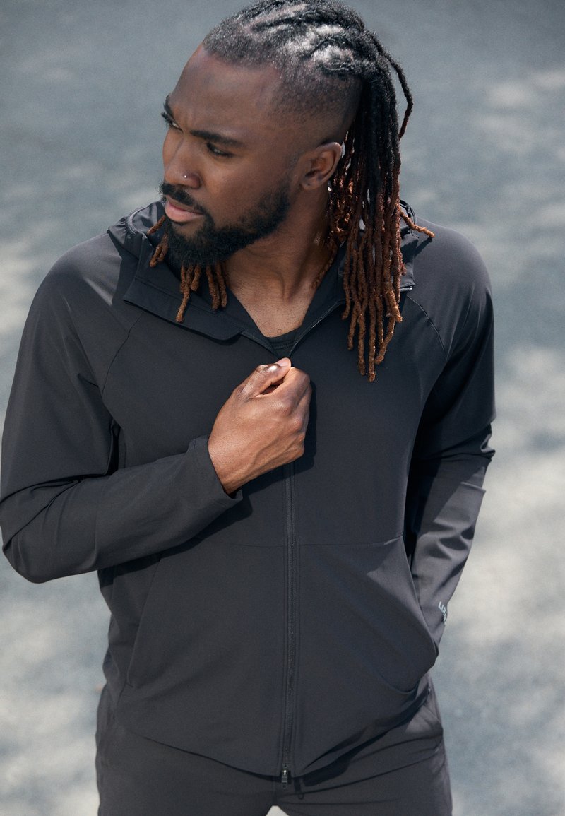 Schwarze, leichte Jacke mit Reißverschluss und Kapuze, taillierte Passform und glatte Textur. Das Modell hält den Kragen und präsentiert die Funktionalität des Kleidungsstücks.