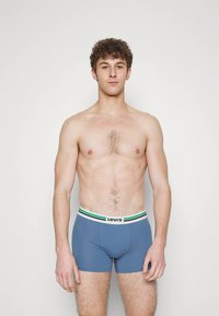 Calções de algodão azuis com uma cintura branca com riscas verdes e pretas, exibidos num modelo sem camisa contra um fundo simples.