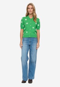 Zelený krátký kardigan s bílými květinovými vzory, zapínáním na knoflíky a vroubkovaným lemem, kombinovaný s světle modrými džíny se širokými nohavicemi.