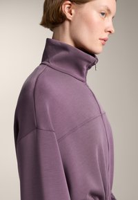 Vijolična zip-up športna jopica z visokim ovratnikom, ki ima gladko teksturo, stranske šive in sproščen kroj.