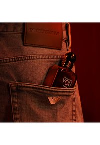 Bruine glazen parfumfles die gedeeltelijk zichtbaar is in een denimzak, gegraveerd etiket met de tekst "Stronger With You," met een metalen logo-accent.