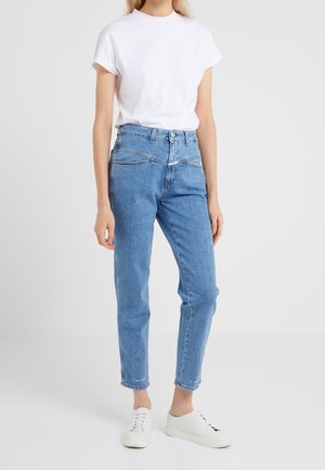 Persoon draagt een wit T-shirt, high-waisted blauwe jeans met asymmetrische voornaad en witte veter sneakers op een effen achtergrond.