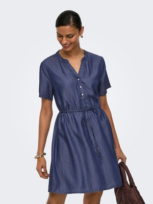 Kobieta ubrana w krótki niebieski denimowy płaszcz z guzikami i paskiem w talii, trzymająca brązową pikowaną torebkę, uśmiechająca się i spoglądająca w dół.