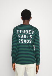 Langarm-T-Shirt in grün und marineblauen Streifen mit weißem Drucktext "ETUDES PARIS 75003" auf dem Rücken. Weicher Baumwollstoff, Rundhalsausschnitt.