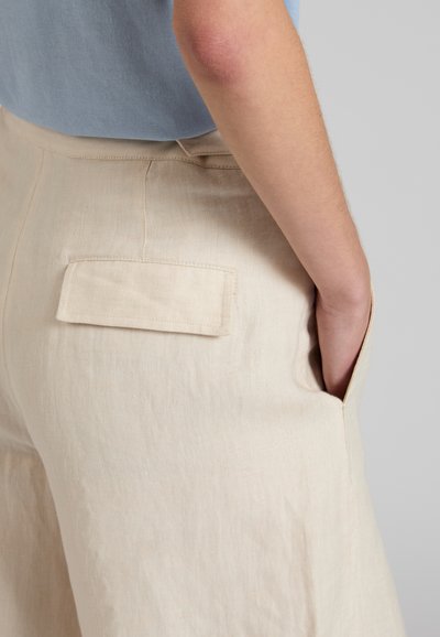 Persona con pantalones de lino beige que tienen una solapa en el bolsillo trasero y la mano en el bolsillo lateral, combinados con una camisa azul claro metida por dentro.