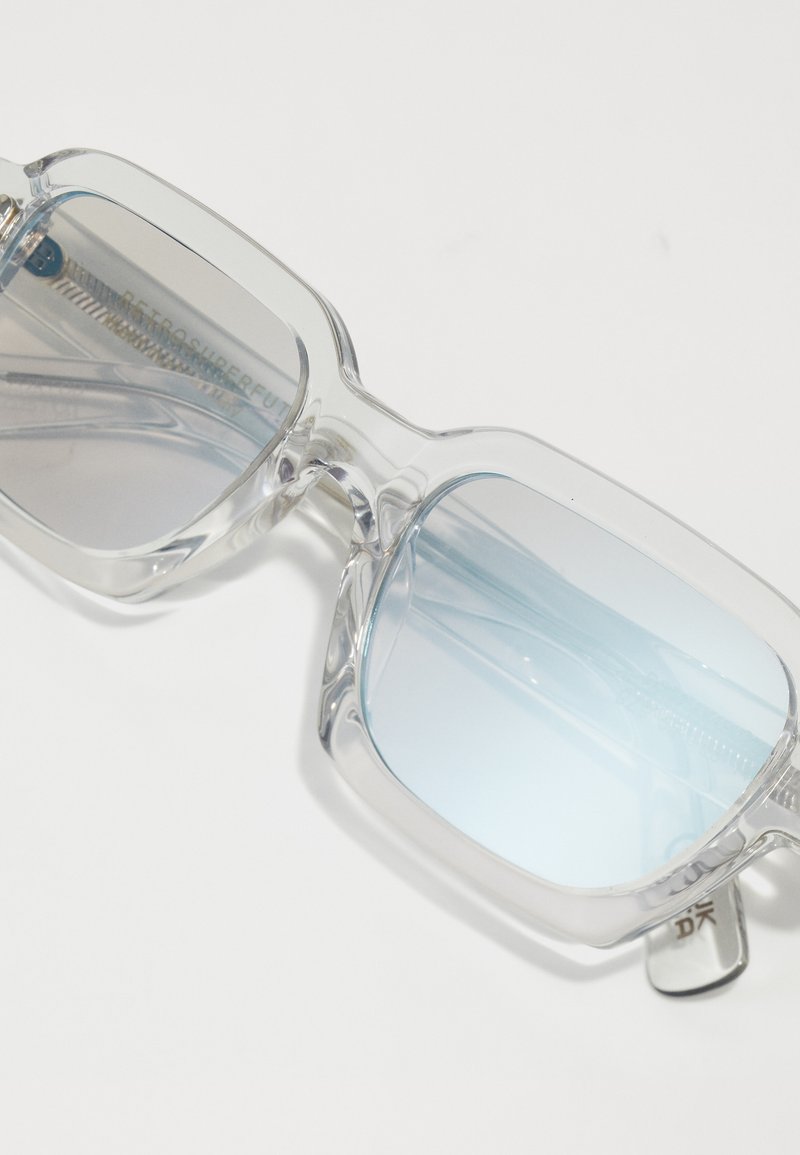 Lunettes de soleil conçues avec une monture en plastique transparent, des verres teintés en bleu, une forme rectangulaire et un branding subtil sur la branche.