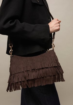 Oseba v črni jakni drži rjavo semiš torbo z resicami in zlatimi okraski, na bež ozadju.