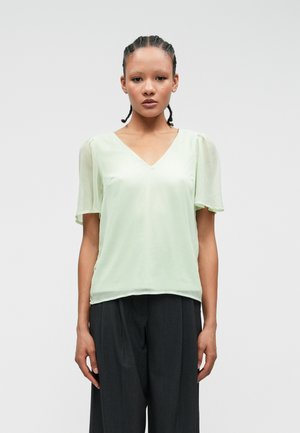 Blouse vert clair, semi-transparente avec un décolleté en V et des manches courtes bouffantes. Texture lisse avec une coupe décontractée. Associée à un pantalon noir ajusté.