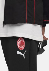 Sort vindjakke med røde accenter over en hvid T-shirt. Sorte sweatpants har et rødt AC Milan-logo og et hvidt Puma-logo.