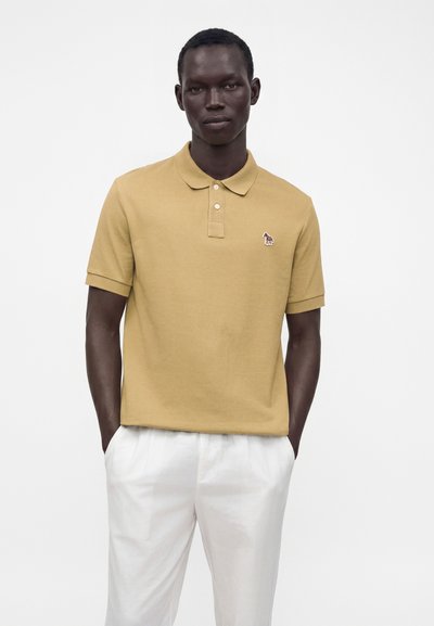 Homme portant un polo beige à manches courtes avec un petit zèbre brodé et un pantalon blanc, debout devant un fond blanc uni.