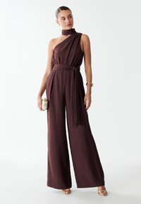 Combinaison une épaule bordeaux avec un tissu texturé et des jambes larges, dotée d'une ceinture nouée et de plis subtils. Accessoirisée avec une petite pochette.