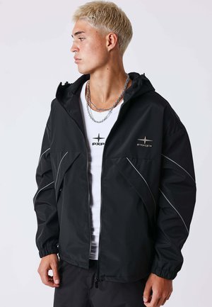 Jeune homme aux cheveux blonds courts portant une veste noire zippée avec des lignes réfléchissantes et un T-shirt blanc avec un logo, regardant sur le côté.