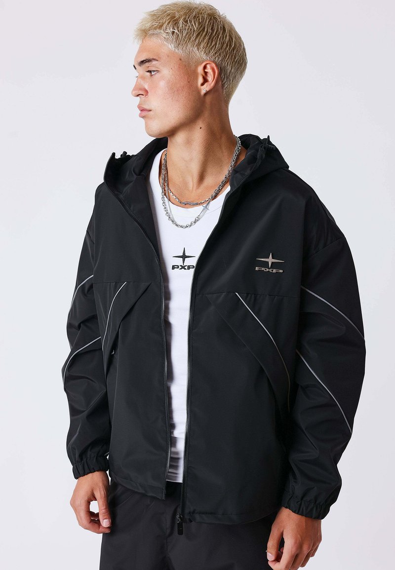 Jeune homme aux cheveux blonds courts portant une veste noire zippée avec des lignes réfléchissantes et un T-shirt blanc avec un logo, regardant sur le côté.