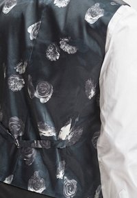 Mężczyzna w czarnym kamizelce w szare kwiatowe wzory, noszący białą koszulę z długim rękawem, widok z tyłu kamizelki.