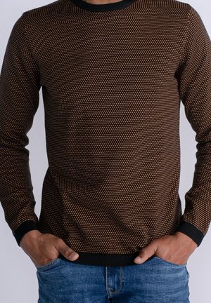 Mand iført en brun og sort mønstret sweater med sorte manchetter og krave, hænderne i lommerne, kombineret med blå jeans.