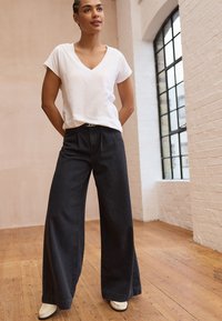 Weißes, kurzärmeliges V-Ausschnitt-T-Shirt kombiniert mit hochtaillierten, weit geschnittenen dunkelblauen Jeans und cremefarbenen Schuhen vor einem Holzboden-Hintergrund.