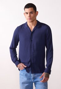 Navyblaues Langarmhemd aus glattem Stoff mit relaxed Kragen, Knopfleiste und taillierter Passform, kombiniert mit hellblauen Jeans.
