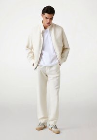 Helle cremefarbene Bomberjacke mit Stehkragen, kombiniert mit passenden cremefarbenen Hosen. Das Modell trägt ein weißes T-Shirt und beige Turnschuhe.