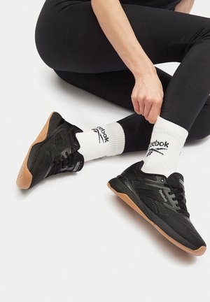 Mustad mustrettis jalanõud, millel on tekstureeritud võrgust pealmine osa, kummist tallad ja pruun aktsent tallal. Kombineeritud valgete logoga sokkidega ja mustade retuusidega.