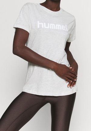 Osoba nosząca jasnoszary krótki rękaw T-shirt z napisem "hummel" i ciemne błyszczące legginsy, ręce splecione na talii.