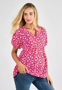 Blusa floreale rosa con maniche a sbuffo corte, scollatura a V e vita arricciata. Presenta fiori bianchi su uno sfondo rosa intenso.