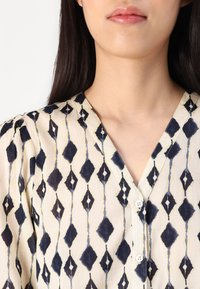 Lightweight fabric blouse with a V-neckline, features a diamond pattern in navy blue on a cream background, button accents at the front.