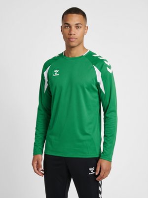 Ung mand iført en grøn sportsskjorte med lange ærmer med hvide accenter og sorte bukser, stående mod en ensfarvet, lys baggrund.