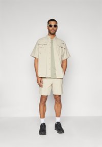 Camicia beige a maniche corte con due tasche, abbinata a pantaloni beige. Indossata con una maglietta verde chiaro e sneakers nere.