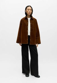 Braune Cordjacke aus Kord mit Knopfleiste und Brusttasche, kombiniert mit schwarzen, weit geschnittenen Hosen und schwarzen Stiefeln.