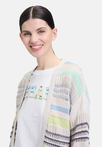 Strickjacke mit offener Maschenstruktur in beigen, blauen, grünen und weißen Streifen. Kombiniert mit einem weißen T-Shirt mit Grafik und mehrfarbigen Buchstaben.