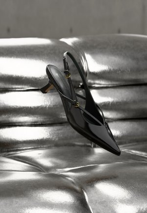 Scarpa slingback in vernice nera con punta affusolata e tacco basso poggiata su una superficie capitonné argento metallico lucido.