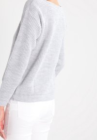 Světle šedý pletený svetr s volným střihem, který má texturovaný vzor a žebrované manžety, v kombinaci s bílými slim-fit kalhotami.