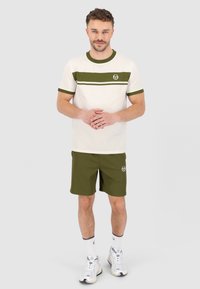 Outfit da uomo con una maglietta color crema con strisce verde oliva e logo, abbinata a pantaloni corti verde oliva e scarpe da ginnastica bianche.