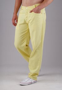 Yellow denim pants with a relaxed fit and textured fabric. Features side pockets and a button closure, paired with white sneakers.