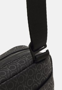Bolso negro de hombro con una superficie texturizada y un patrón de logo repetido, equipado con una correa negra elegante y un clip de metal.