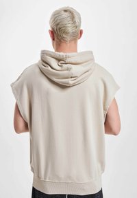 Beige ärmelloser Hoodie mit weicher Textur, ausgestattet mit einer großen Kapuze und geripptem Saum, getragen von einer Person, die sich abwendet.