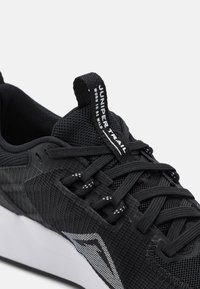 Scarpa sportiva nera con tomaia in mesh strutturato, lacci audaci e logo con bordo bianco sulla linguetta e sul lato, con suola bianca.