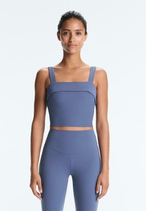 Kobieta stojąca przodem, ubrana w niebieski prążkowany krótki top z szerokimi ramiączkami oraz dopasowane legginsy z wysokim stanem na białym tle.