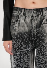 Close-up of waist and hand wearing black jeans with silver studded patterns fading upward from thighs to waist.