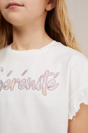 Bambino con lunghi capelli biondi che indossa una camicia bianca con la parola "Sérénité" ricamata in colori pastello e maniche corte arricciate.