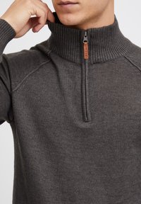 Ciemnoszary dzianinowy pulower z wysokim prążkowanym kołnierzem i zamkiem błyskawicznym na pół. Z ozdobnym skórzanym zawieszką i subtelnymi detalami tekstury.