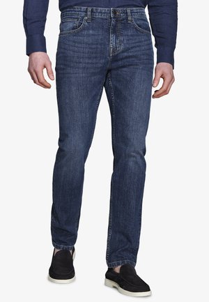 Mężczyzna ubrany w ciemnoniebieskie dopasowane dżinsy, ciemnoniebieską koszulę z długim rękawem oraz czarne wsuwane buty z białymi podeszwami, stojący na jednolitym tle.